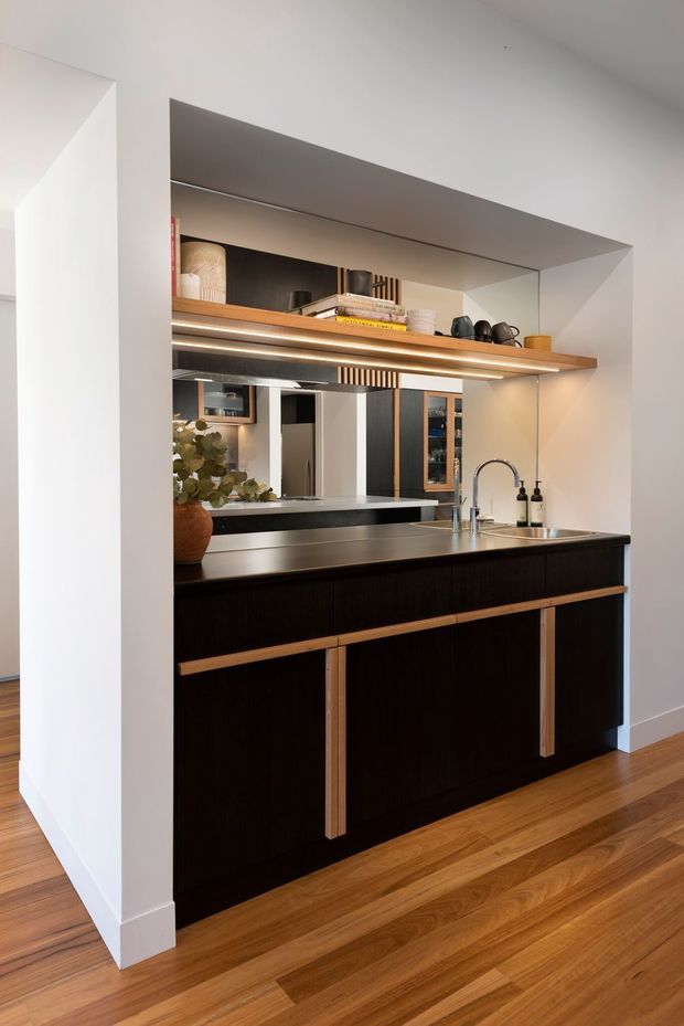 A compact, built-in kitchen or kitchenette with a dark timber island and cabinetry, slim light-wood handles, a dark benchtop, stainless-steel sink and tap, and a mirrored splashback. An illuminated open timber shelf displays books and decorative objects above the work area. Warm timber flooring and white walls create a refined, contemporary interior.
