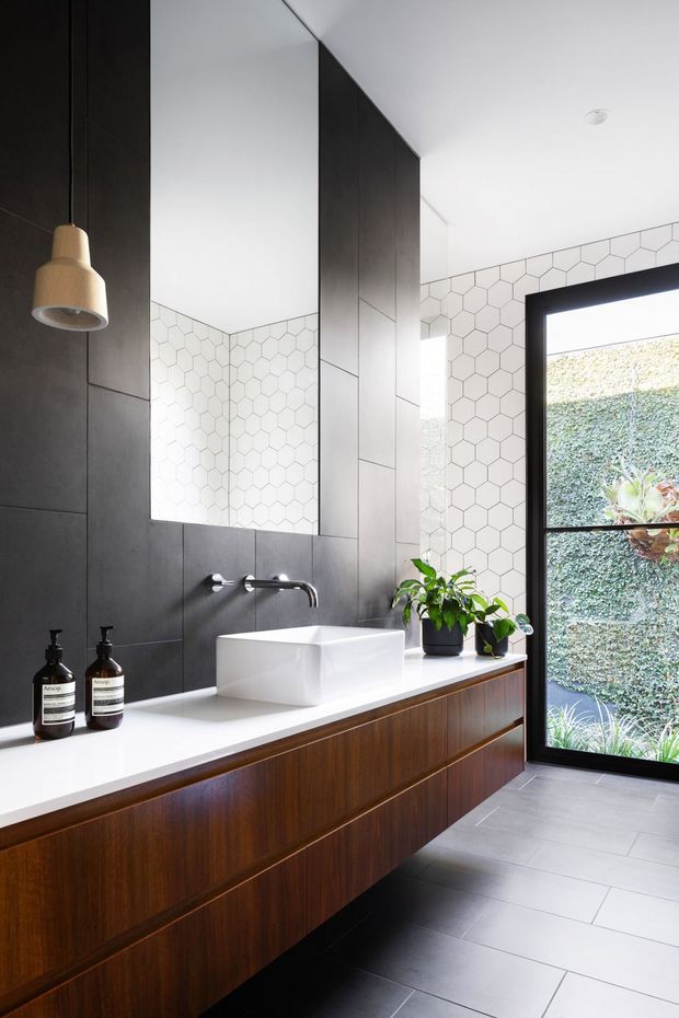 A contemporary bathroom featuring a long floating timber vanity with a white solid-surface benchtop, rectangular vessel basin, wall-mounted chrome tapware, and a large frameless mirror. The walls combine dark large-format tiles with white hexagonal tiles, while grey floor tiles and black-framed glazing open to a planted exterior courtyard. A pendant light and potted plants add visual interest.
