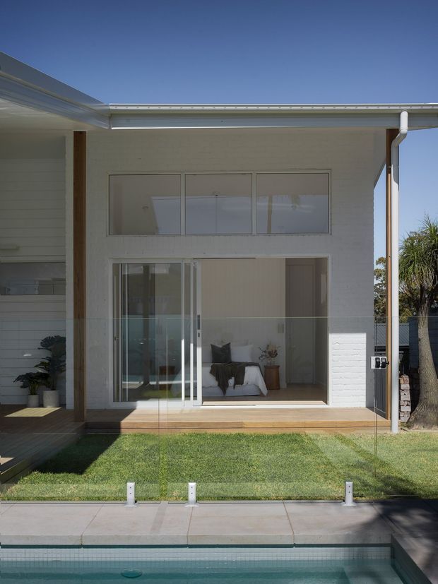Exterior view of a contemporary two-storey residential extension or rear facade opening onto a swimming pool. The white horizontal-clad facade has broad glazing, a sliding glass door, timber posts, a shallow roof overhang, and a lawn terrace between the house and pool. A bedroom or living area is visible through the glazing.