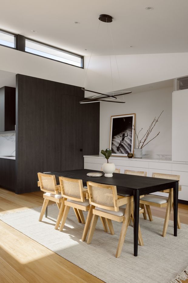 A refined open-plan dining area with a long black dining table and six light timber cane-back chairs positioned on a large neutral rug. The room features pale timber flooring, white walls and ceiling, extensive dark timber wall panelling and concealed cabinetry, high clerestory windows, and a sculptural suspended linear pendant. Minimal artwork, a vase, and branches provide restrained decoration.