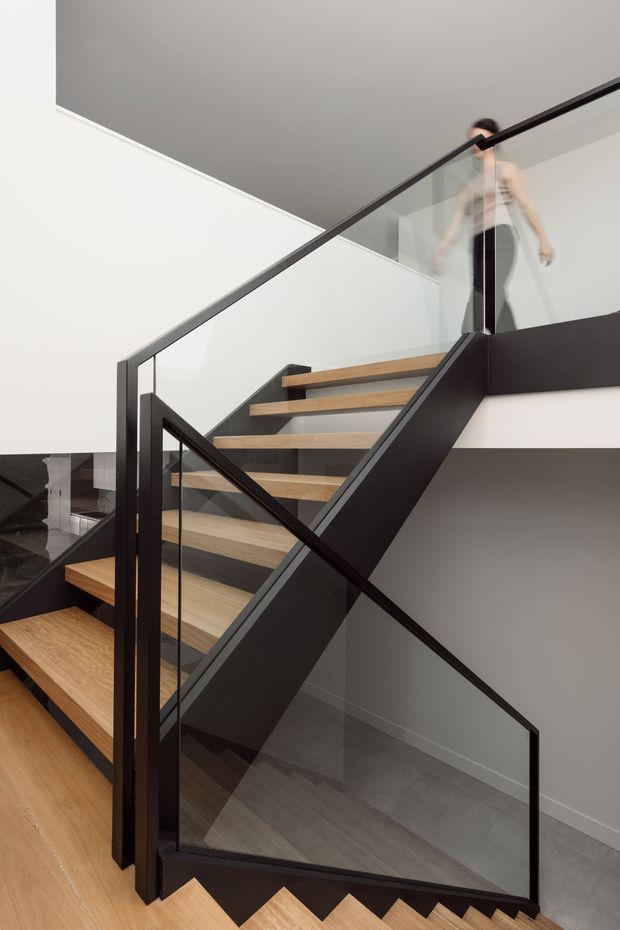 Contemporary stairway with open timber treads, black-painted steel stringers and balustrade, and clear glass panels. The staircase connects multiple levels in a bright minimalist interior with white walls, a grey ceiling, and contrasting dark structural elements.