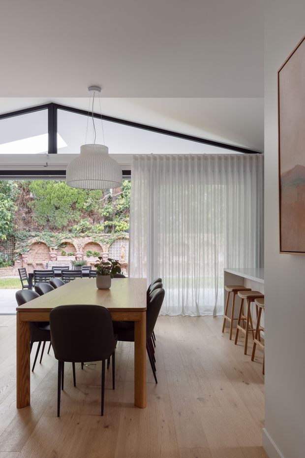 A refined dining area in a contemporary home, with a large timber dining table, dark upholstered chairs, a sculptural pendant light, sheer curtains, and pale timber flooring. Full-height glazing opens to a leafy brick courtyard, while a minimalist white island or bar with timber stools is visible to the right. Dark-framed angular glazing and warm natural materials create a calm indoor-outdoor connection.