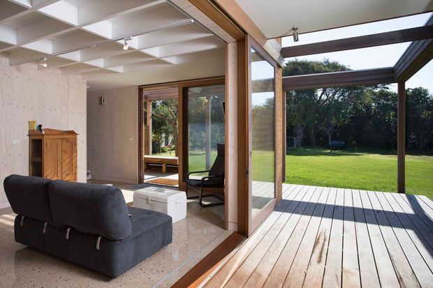 A bright contemporary living room opening directly onto a broad timber deck and large landscaped lawn through extensive timber-framed glazing. The room includes a dark upholstered sofa, a wood cabinet, pale walls, a polished light terrazzo-like floor, and a distinctive white coffered ceiling. The covered deck uses exposed dark timber beams and posts, creating a strong indoor–outdoor connection.