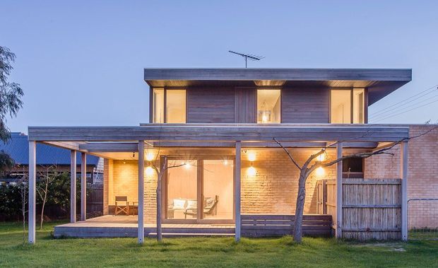 Twilight exterior view of a contemporary two-storey residence with a strong horizontal flat roof, warm timber cladding, extensive glazing, and a covered outdoor terrace or patio. The lower level combines pale brick walls with timber screening and slender posts, while the upper level features recessed illuminated rooms and a broad cantilevered roof edge. A lawn and mature tree sit in the foreground.