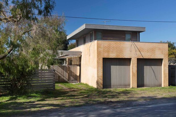 Exterior street view of a compact two-storey residential building with a prominent double garage, pale warm brick walls, corrugated garage doors, timber screening and fencing, and a flat roof with broad overhanging eaves. Mature trees and a driveway frame the property.