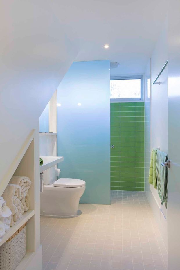 Bright compact bathroom with a white toilet and wall-mounted basin, pale tiled flooring, a vivid green tiled shower wall, white walls, built-in open towel shelving, and a small frosted window. The space has a clean, playful contemporary fit-out with pastel aqua and green accents.