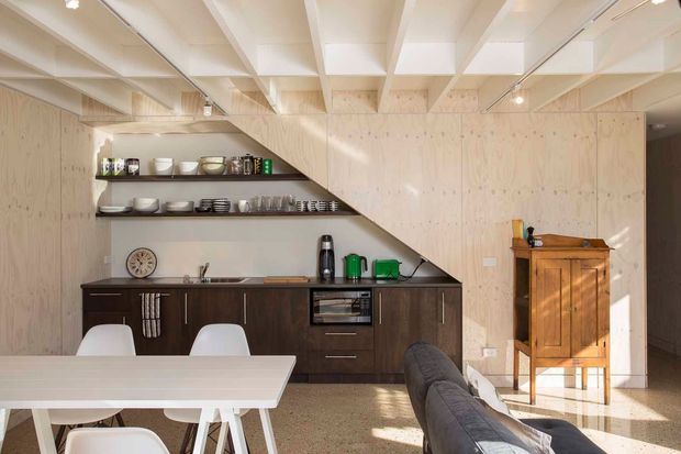Bright open-plan kitchen and dining area with a compact dark-wood galley kitchen beneath a sculptural timber stair or overhead structure. The space features pale plywood wall and ceiling surfaces, open shelving displaying crockery, integrated appliances, a light dining table with white chairs, and a grey sofa in the foreground. Strong sunlight creates a warm, contemporary atmosphere.