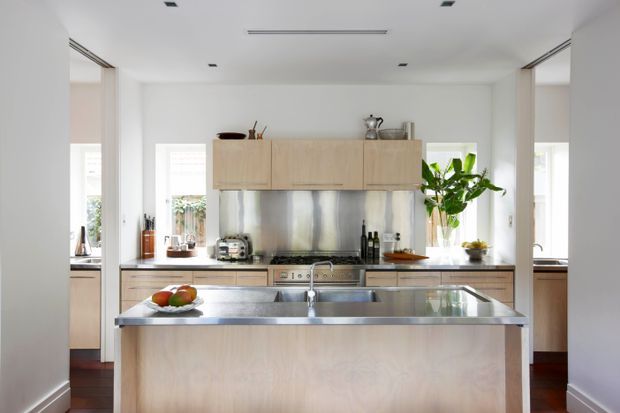 Bright contemporary kitchen viewed directly toward a large central island. The room features pale timber cabinetry, a stainless-steel benchtop and splashback, integrated appliances, white walls, recessed ceiling lights, and abundant natural light from multiple windows. A leafy indoor plant and minimal countertop items add warmth to the otherwise clean, refined space.
