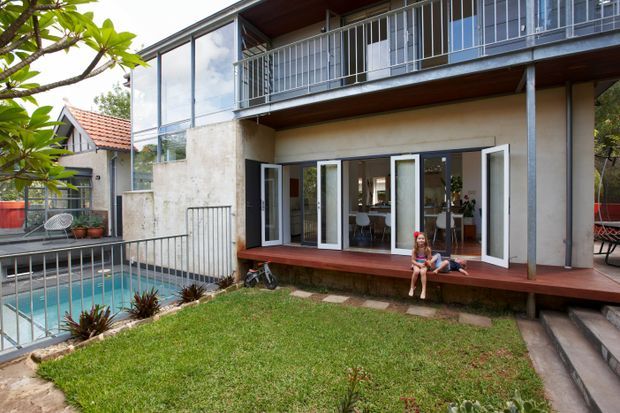 Exterior view of a two-storey contemporary residential building with a concrete lower facade, extensive white-framed glazing, open upper-level balcony, and dark timber soffits and decking. A fenced swimming pool, lawn, garden planting, and outdoor seating are visible around the house.