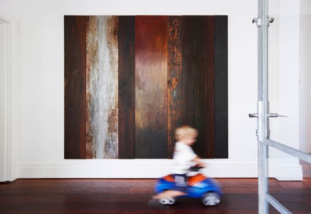 A bright, sparsely furnished interior with white walls and trim, dark timber flooring, and a large vertically panelled abstract wall feature made from varied weathered and stained timber boards. A blurred child on a ride-on toy crosses the foreground, while a partially visible framed glass-and-metal doorway or partition appears at the right.
