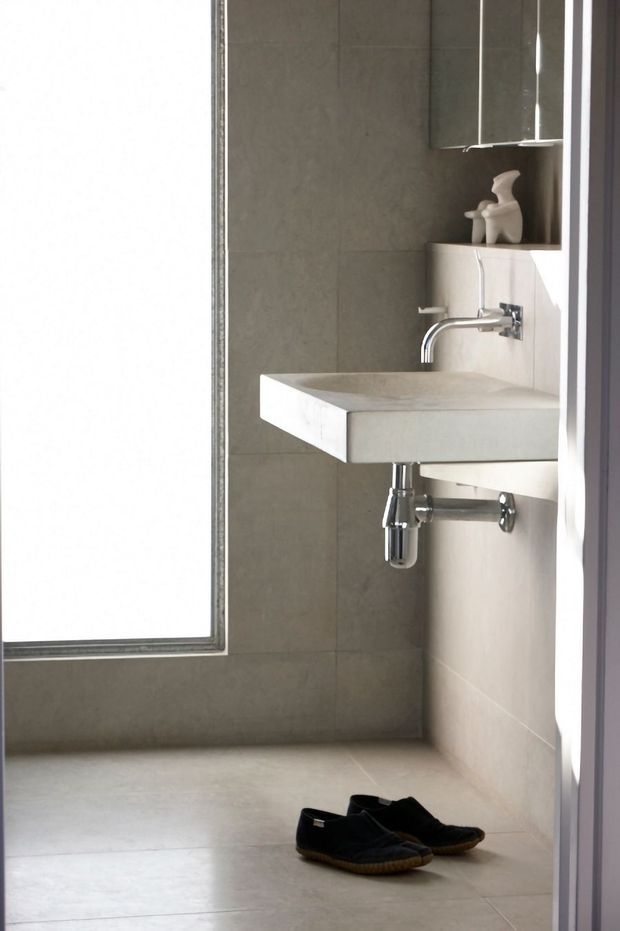 A compact contemporary bathroom with a wall-mounted rectangular concrete or stone-look basin, exposed chrome plumbing, wall-mounted chrome faucet, pale grey-beige large-format wall and floor tiles, and a mirrored cabinet above the vanity. Natural light enters from a tall window on the left, creating a clean, restrained atmosphere.