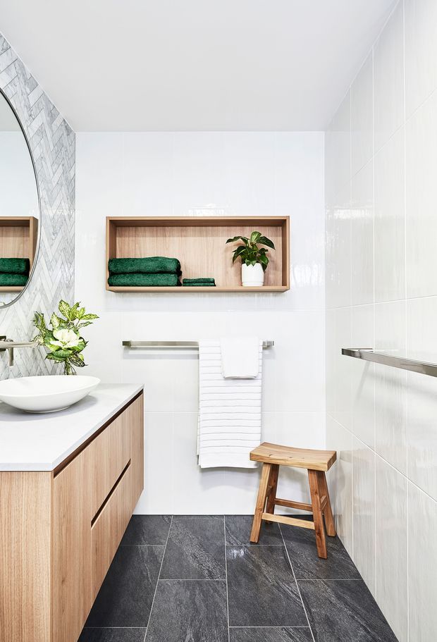 Bright, contemporary bathroom with a floating light-wood vanity, white vessel basin, large round mirror, wall-mounted timber display niche holding folded green towels and a plant, white wall tiles, patterned feature tiles around the mirror, and dark charcoal floor tiles. A timber stool and white towels add warmth and contrast.
