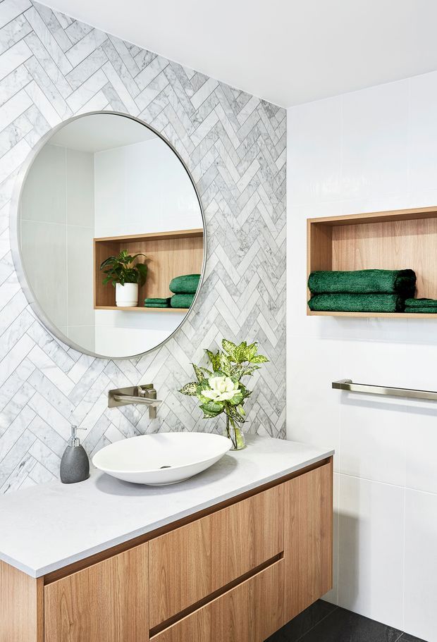 Bright contemporary bathroom vanity featuring a floating timber-grain cabinet, white stone benchtop, vessel basin, wall-mounted brushed metal tapware, large round mirror, and recessed timber display niches. The main wall is finished in a white and grey marble-look herringbone tile, with white tiled walls and a dark floor creating contrast.