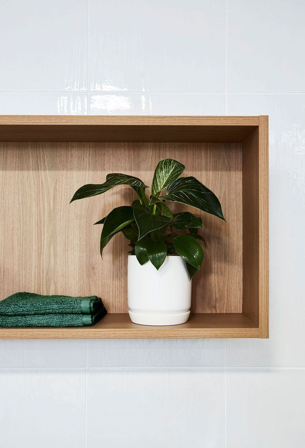 Close-up of a minimalist built-in timber wall niche or open shelving unit set into glossy white tiled walls. The shelf contains a white ceramic planter with a leafy green plant and folded dark green towels, creating a simple, natural bathroom storage display.