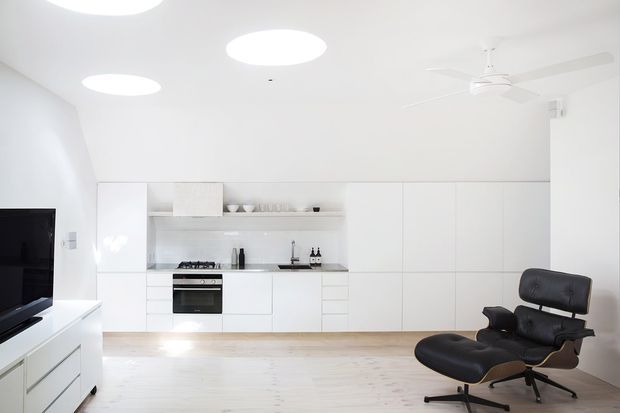 Bright minimalist open-plan living area with a compact white kitchen integrated into a wall of full-height cabinetry. The room has pale timber flooring, white walls and ceiling, circular recessed ceiling lights, a white ceiling fan, and a black leather lounge chair. A television and low white media cabinetry are visible at the left.