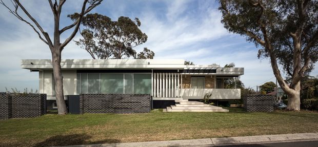 A low, single-storey contemporary house viewed from the street, set behind a broad lawn and mature eucalyptus trees. The facade combines pale rendered walls, extensive horizontal glazing, a flat roof, dark brick privacy walls, and a sheltered entry reached by broad steps. Vertical timber or metal screening defines the central entrance zone, while a covered carport-like structure extends at the right.
