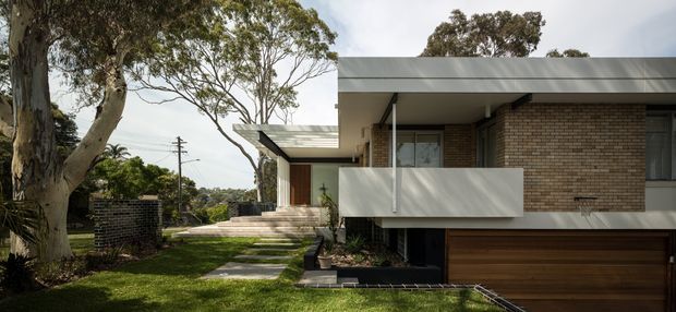 Exterior view of a contemporary multi-level brick residence set among mature trees and landscaped grounds. The house features a broad low-pitched roof, warm brown brickwork, white rendered or metal projecting balcony elements, dark-framed glazing, stepped entry paths, and an integrated timber-toned garage door.