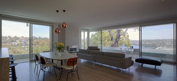 A bright open-plan contemporary living and dining area with expansive corner glazing overlooking a leafy urban waterfront setting. The room features warm timber flooring, a neutral grey sofa, a light dining table with mixed chairs, minimalist built-in cabinetry, and three copper pendant lights. Large sliding glass doors open toward a balcony or terrace, creating an airy indoor-outdoor connection.