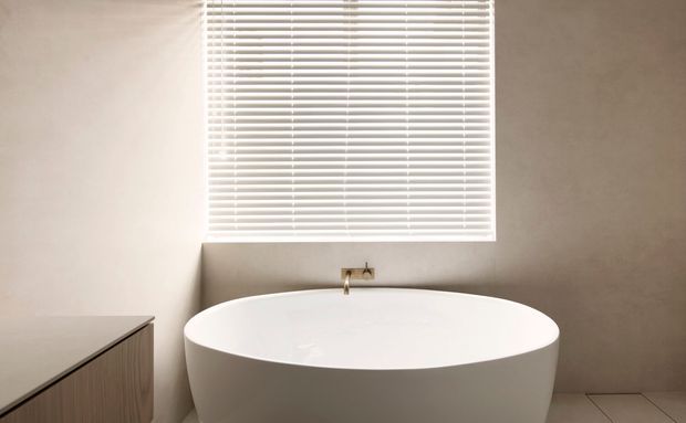 Minimalist bathroom featuring a freestanding white oval bathtub beneath a large window with white horizontal blinds. A compact timber-toned vanity with a pale benchtop is visible at left, while the walls and tiled floor are finished in warm neutral tones. Brass or gold-toned wall-mounted bath fittings add a subtle accent.