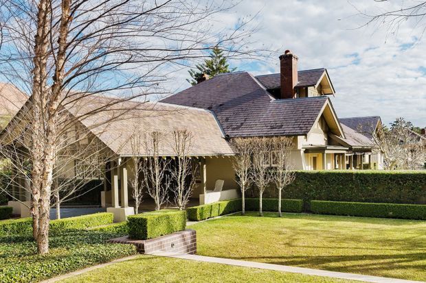 Exterior view of a large traditional residence with multiple pitched gabled roof sections, dark shingle-like roofing, pale rendered walls, a prominent brick chimney, covered entry or veranda, and carefully manicured formal hedges and lawn.