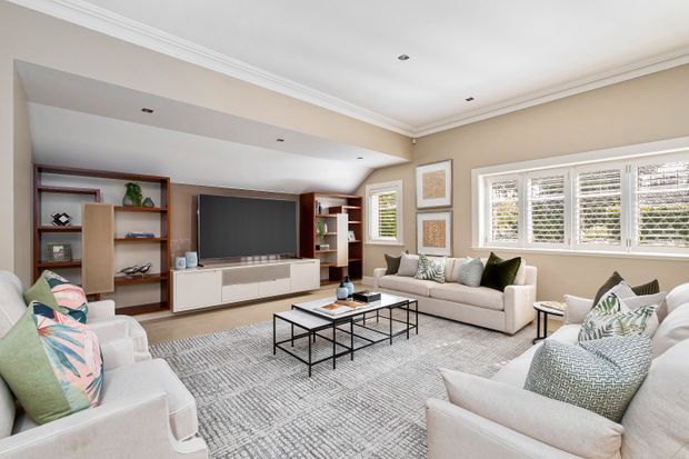 Bright, spacious living room with a neutral upholstered sofa and armchairs arranged around nested black metal-and-glass coffee tables. Built-in timber-toned shelving and cabinetry frame a large wall-mounted television, while plantation shutters, framed artwork, layered cushions, a textured area rug, and decorative accessories create a refined contemporary family-room setting. Crown moulding and recessed ceiling lights add traditional architectural detailing.