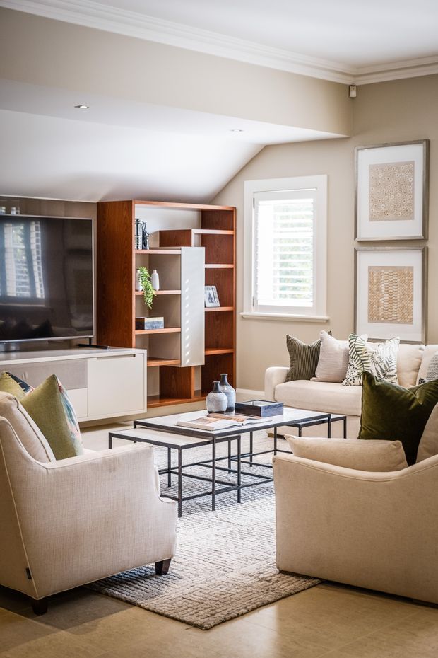 A bright, refined living room with a neutral beige and cream palette. The room features upholstered armchairs and a sofa arranged around nested black metal-and-glass coffee tables, layered rugs, framed artwork, and a built-in timber entertainment and shelving unit with white cabinetry. A shuttered window provides daylight, while detailed ceiling cornices and recessed lighting add a classic architectural finish.