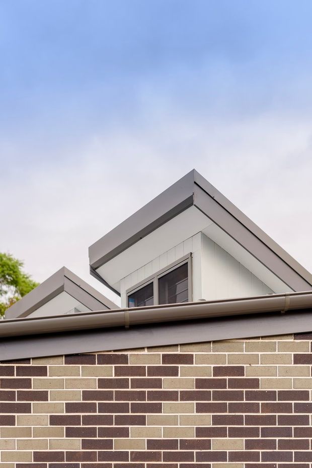 Close-up exterior view of a contemporary residential building, focused on overlapping pitched roof forms, box-like upper-level cladding, dark-framed windows, gutters, and a patterned multi-tone brick wall beneath. The image is photographed from below against a pale blue sky.