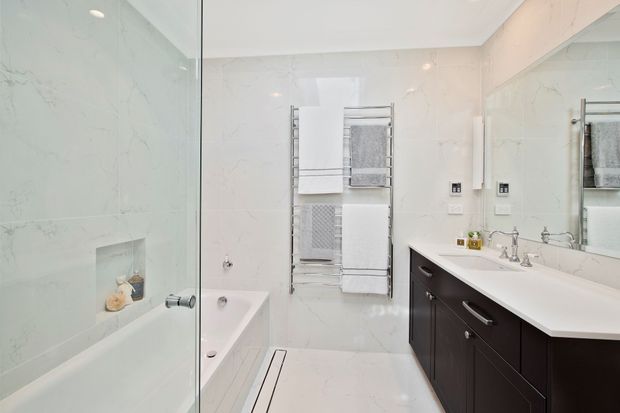 Bright contemporary bathroom with large white marble-look wall and floor tiles, a frameless glass shower enclosure, built-in bathtub, heated towel rail with folded towels, and a dark wood vanity with a white countertop and integrated basin. A large mirror spans the vanity wall.