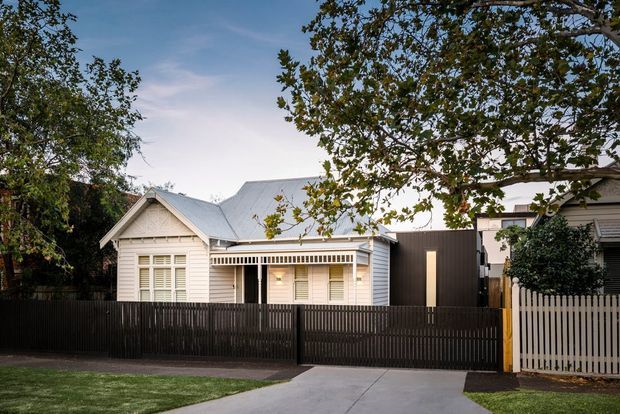Street-facing view of a renovated single-storey home combining a white weatherboard cottage with a darker contemporary extension. The facade includes a pitched metal roof, decorative verandah detailing, shuttered windows, and contrasting timber picket and slatted fencing, with a concrete driveway and mature trees framing the property.