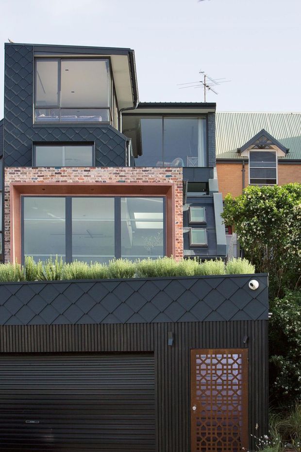 Exterior street-facing view of a multi-level contemporary residence, featuring dark diamond-pattern cladding, black vertical wall paneling, extensive aluminium-framed glazing, a brick-framed upper window, and an integrated garage with a decorative metal pedestrian door. Landscaping and neighbouring buildings are visible around the facade.