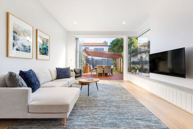 Bright contemporary living room with a large light-coloured sectional sofa, blue cushions, framed artwork, a textured area rug, and a wall-mounted television set into white built-in joinery. Full-width glazed doors open directly onto a timber-decked outdoor dining area with a pergola, creating a strong indoor-outdoor connection.