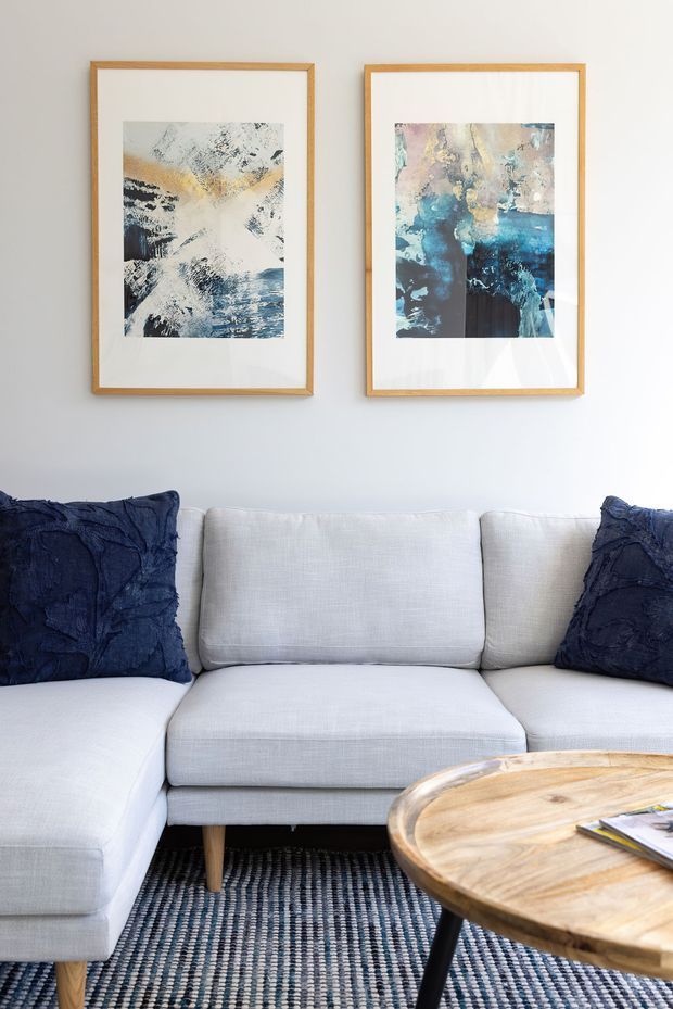 Bright, calm living room vignette featuring a large light-grey sectional sofa with navy textured cushions, a round natural timber coffee table, and a blue-grey woven rug. Two abstract blue, cream and ochre artworks in slim natural timber frames hang on a plain white wall.