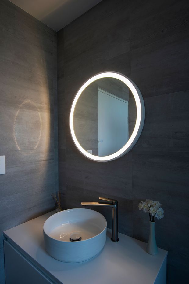 A compact, moody contemporary bathroom vanity viewed close-up. The walls are clad in dark grey large-format textured panels, with a white countertop supporting a round vessel basin and a dark metal single-lever faucet. A large circular mirror with integrated LED illumination forms the main feature above the vanity. Minimal decorative elements include a slim vase with flowers and a small reed arrangement.