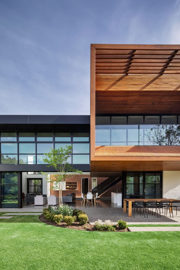 A contemporary two-storey residential building viewed from the landscaped rear garden. The facade combines expansive black-framed glazing, dark horizontal cladding, exposed red brick, and a prominent warm timber-lined upper volume with a slatted pergola roof. The ground level opens onto a paved outdoor dining and lounge area, with lawn and planted garden beds in the foreground. An interior living area, dining area, and stair are visible through the glazing.