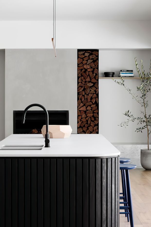 A refined contemporary kitchen centred on a large island with a white benchtop, black gooseneck tap, integrated cooktop and dark vertical timber slat cabinetry. Behind the island is a minimalist white wall with a black fireplace or recessed feature containing stacked firewood, open shelving with books, and a sculptural olive tree. Blue stools sit at the island, while pale timber flooring and a restrained neutral palette create a clean, modern atmosphere.