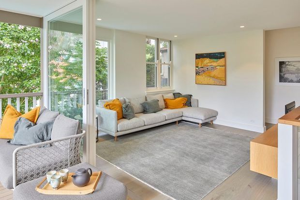 Bright contemporary living room with a large light-grey sectional sofa, grey area rug, timber flooring, and warm yellow-orange cushions. Floor-to-ceiling glazing opens onto a leafy balcony or terrace, bringing abundant natural light into the room. A timber media console and framed artwork add understated character.