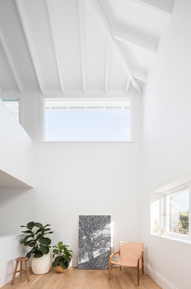 Bright, minimalist white interior with a dramatic high pitched exposed-beam ceiling and clerestory-style glazing. The sunlit room features pale timber flooring, white walls, a large leafy plant, a small wooden stool, an upholstered wooden armchair, and a monochrome artwork leaning against the wall. The space feels airy and serene, with strong natural light and a double-height volume.