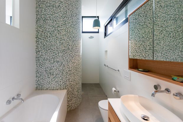 Bright contemporary bathroom with a freestanding-style white bathtub, curved mosaic-tiled shower enclosure, wall-mounted toilet, and double white vessel basins set into timber cabinetry. A large mirrored cabinet and high clerestory windows add light and reflect the room.