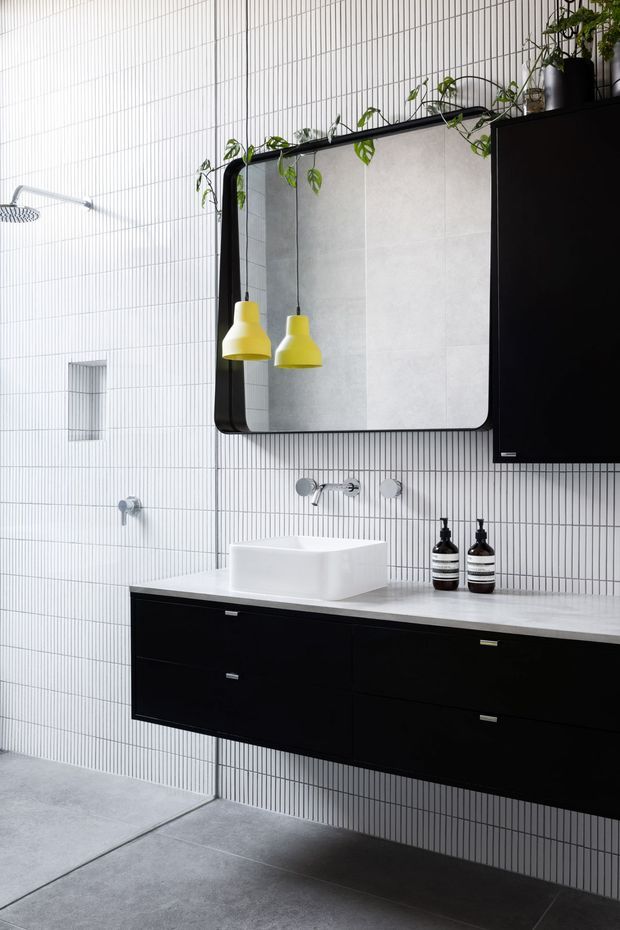 A contemporary bathroom featuring a floating black vanity with integrated drawers, a white rectangular vessel basin, wall-mounted chrome tapware, and a large black-framed mirror. The walls are clad in narrow vertically striped white tiles, complemented by large grey floor tiles. Yellow pendant lights hang in front of the mirror, with trailing plants adding a small organic accent. A black wall cabinet and monochrome bathroom accessories complete the refined, high-contrast scheme.
