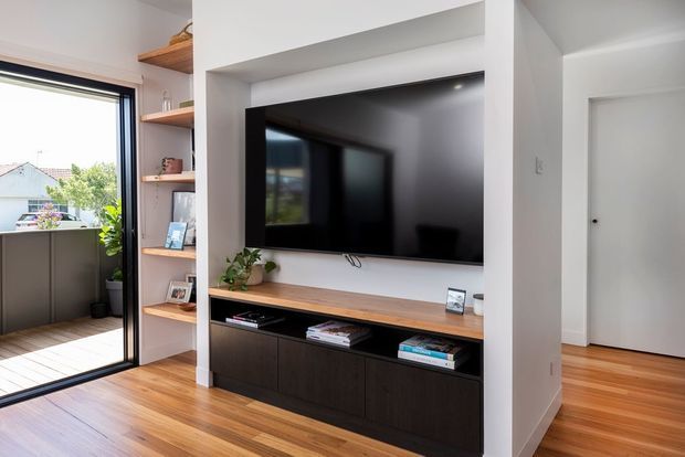 Bright contemporary living room media wall with a large black television set into a white built-in niche, a timber display shelf and benchtop, and dark built-in cabinetry below. Warm timber flooring continues toward a white hallway and closed interior door. A large glazed opening provides daylight and views outside.