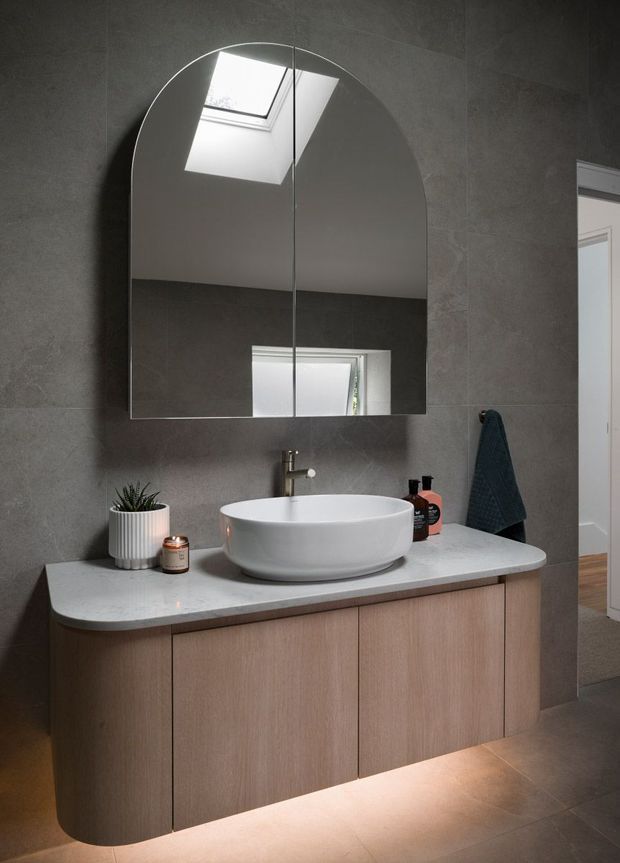 Contemporary bathroom vanity with a floating, rounded-edge timber-look cabinet, pale stone benchtop, white vessel basin and brushed metal tapware. A large arched mirror reflects a skylight and window, while grey wall and floor tiles create a restrained, modern palette. Under-vanity lighting adds a warm glow.
