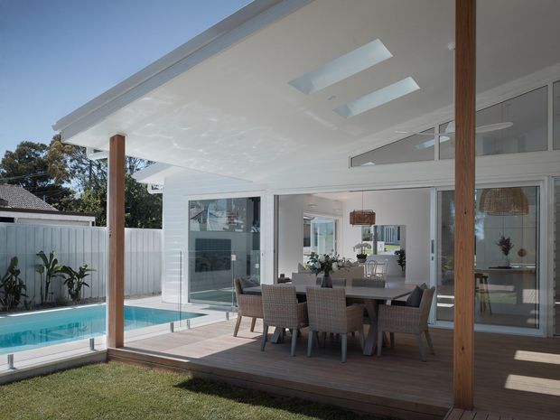 Bright modern covered outdoor entertaining area adjoining a glazed living space, with a timber deck, dining table and chairs, timber posts, skylights, and a swimming pool beside the patio. White walls, extensive sliding glass doors, and clerestory glazing create an airy indoor-outdoor connection.