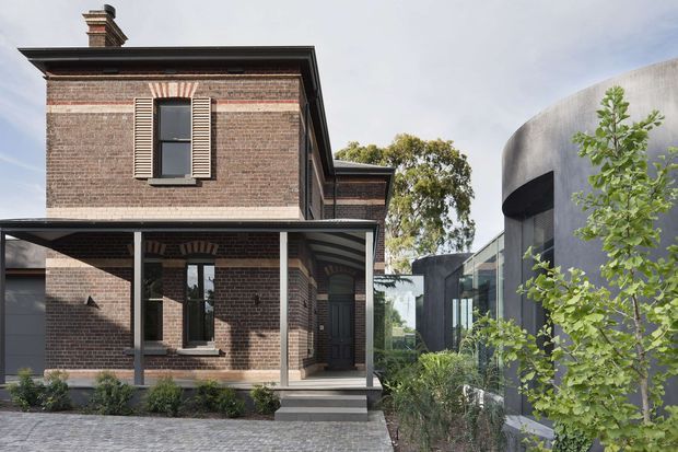 Exterior architectural photograph showing a restored two-storey red-brick heritage house with decorative brick detailing, a covered front veranda, timber shutters, dark trim, and a traditional chimney. A contrasting contemporary addition with curved concrete walls and extensive glazing is visible to the right, connected through landscaped grounds and a paved forecourt.
