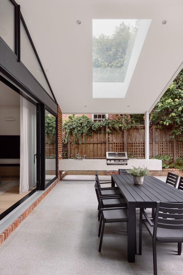 A covered outdoor dining terrace adjoining a contemporary home, with a large black dining table and chairs on a pale textured floor. The terrace has a white ceiling with a large skylight, slim white columns, a built-in white barbecue bench, exposed brick detailing, dark-framed sliding glazing, and a timber garden fence with climbing greenery beyond.
