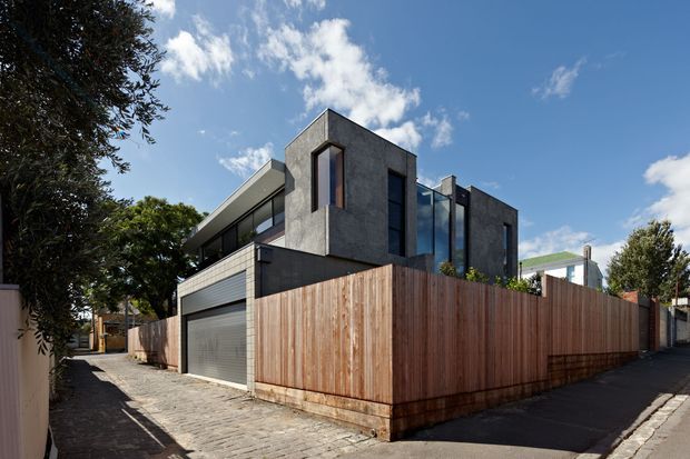 Street-level exterior view of a contemporary multi-storey urban residence. The house has dark textured masonry or rendered concrete volumes, extensive vertical glazing, a projecting upper-level balcony, a concealed garage, and a tall vertical timber privacy fence. It is set along a paved urban lane with neighbouring homes and mature trees.