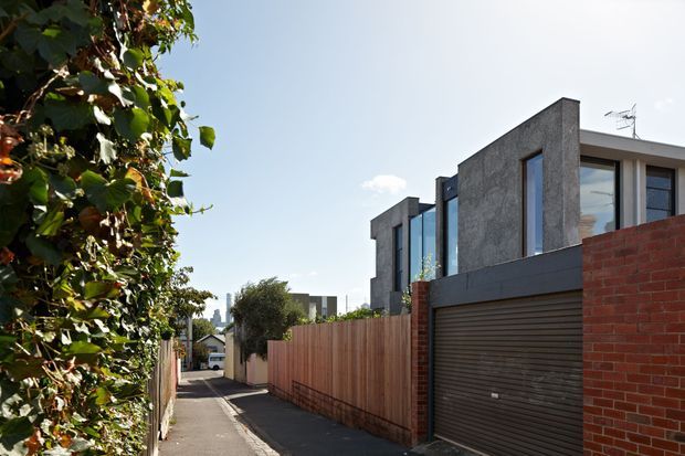 Street-level view along a narrow urban residential laneway, with a contemporary multi-storey house occupying the right side. The home features dark textured masonry or concrete walls, large vertical windows, a flat roofline, a dark sectional garage door, and a red brick boundary wall. Timber fencing and dense greenery line the laneway, with additional modern residences visible in the background.
