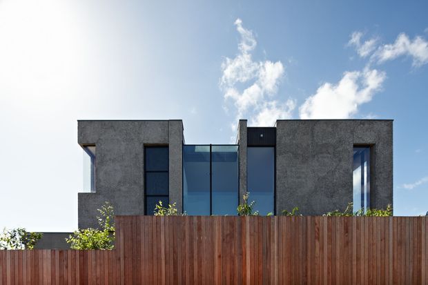 Front exterior view of a contemporary two-storey residence behind a tall vertical timber privacy fence. The building has strongly rectilinear volumes, dark textured rendered walls, narrow vertical windows, and a central floor-to-ceiling glazed section. A bright blue sky is visible above the roofline.