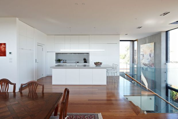 Bright open-plan kitchen and dining area with extensive white built-in cabinetry, a central island, stainless-steel appliances, timber flooring, and a traditional timber dining table in the foreground. Full-height glazing and a glass balustrade overlook an exterior space, while recessed ceiling lights and clean-lined joinery create a contemporary interior.