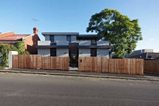 Street-facing view of a contemporary multi-level urban residence with a flat roof, box-like massing, dark-framed glazing, pale rendered walls, projecting balconies, and a timber slat privacy fence. A mature tree partially screens the facade, while older brick and tiled-roof homes are visible on either side.