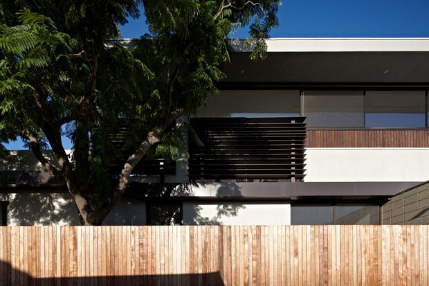 Exterior view of a contemporary multi-level residence with a flat roof, white rendered walls, extensive glazing, dark horizontal timber privacy screens, and warm vertical timber fencing. Mature vegetation partially screens the facade.